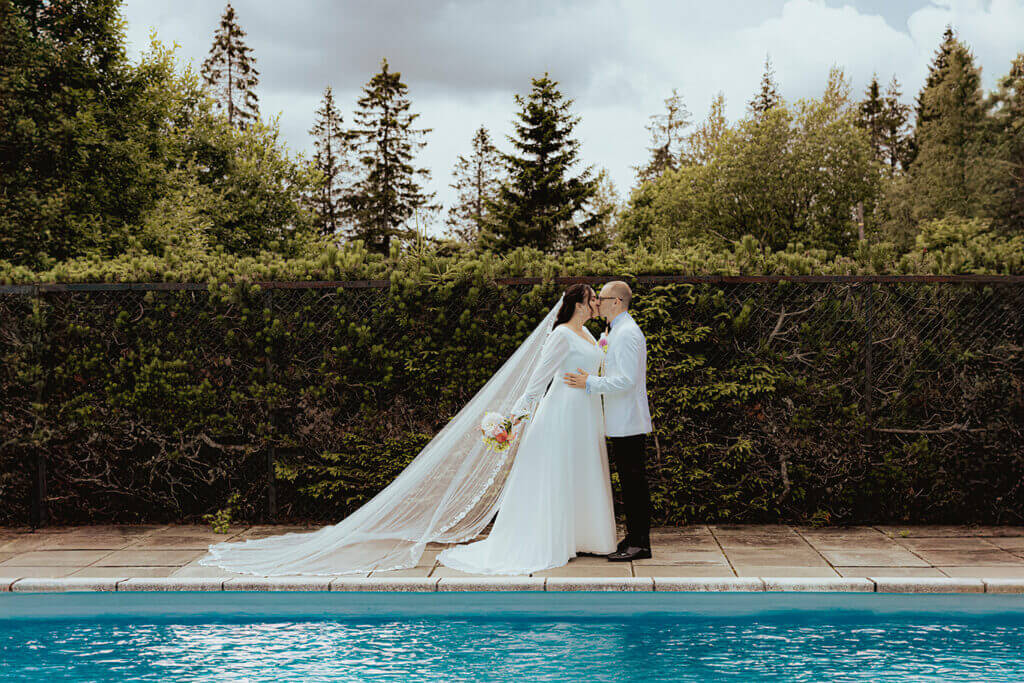 Brudepar deler et kyss ved et basseng, omgitt av frodig natur. Et romantisk øyeblikk fanget av Anna O. Brambilla Photography, perfekt for en artikkel om bryllupsplanlegging og kjørelpan for bryllup på Oslo Rådhus.