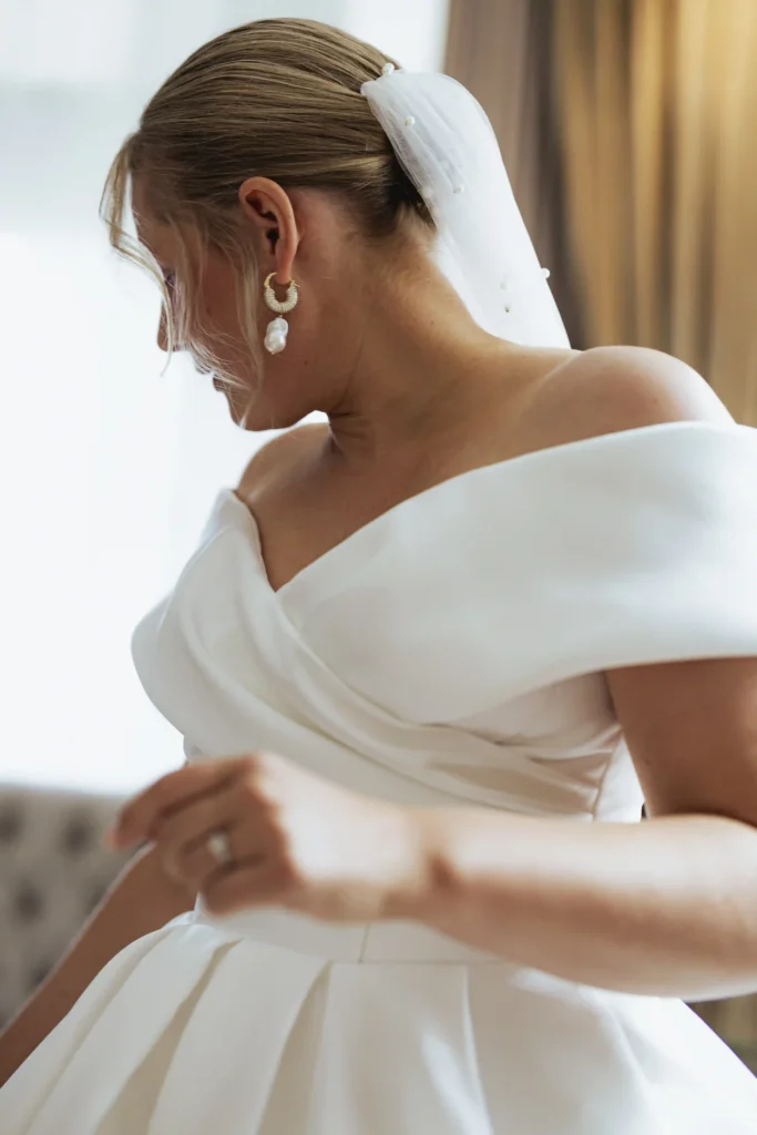 En vakker brud i en hvit off-the-shoulder brudekjole og perleøredobber justerer kjolen sin før sitt norsk-italienske bryllup. Fotografert av Anna O. Brambilla. A beautiful bride in a white off-the-shoulder wedding dress and pearl earrings adjusts her gown before her Norwegian-Italian wedding. Captured by Anna O. Brambilla.