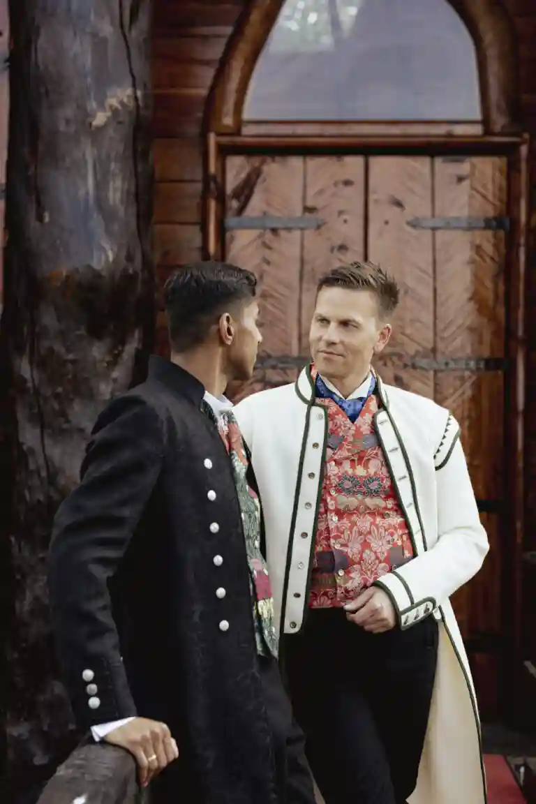 Bryllupsfotograf Anna O. Brambilla fotograferer to brudgommer i vakre, tradisjonelle bunader, mens de ser kjærlig på hverandre foran en stemningsfull trekirke.