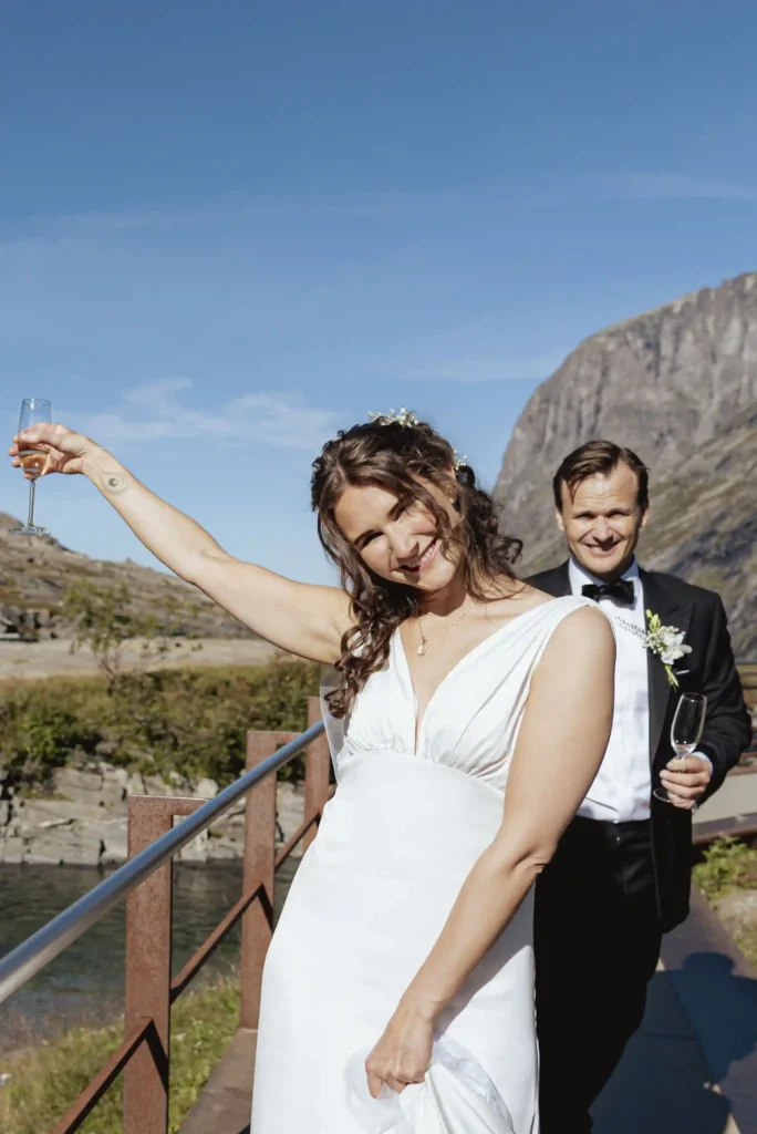 Brudepar som feirer med champagne på Trollstigen på en solrik bryllupsdag. Bruden smiler og løfter glasset, mens brudgommen står bak og smiler, med dramatiske fjell i bakgrunnen. Fotografert av Anna O. Brambilla