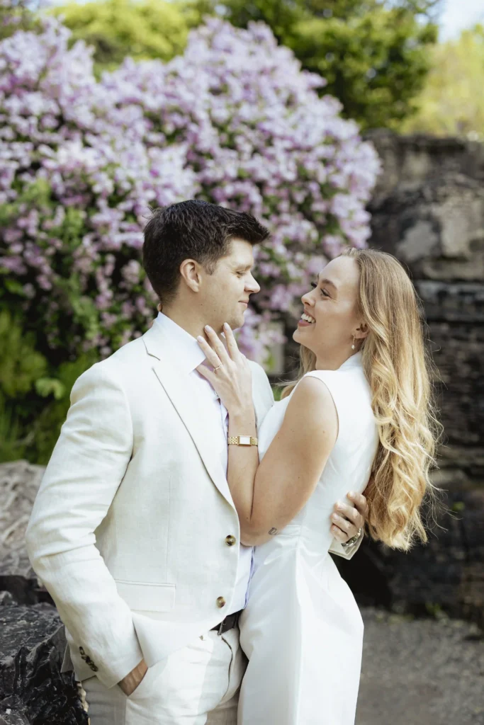 A joyful couple dressed in white sharing a romantic moment in front of blooming lilac flowers. The woman smiles up at her partner while gently touching his chest, and they both look lovingly at each other. Photo by Anna O. Brambilla.