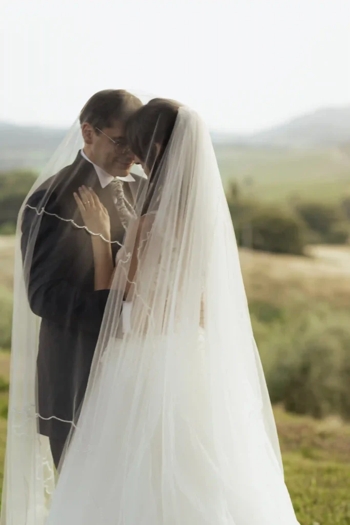 Brudepar står tett omslynget under brudesløret med utsikt over det toskanske landskapet. Et intimt øyeblikk fra et romantisk bryllup i Toscana. Foto av bryllupsfotograf Anna O. Brambilla