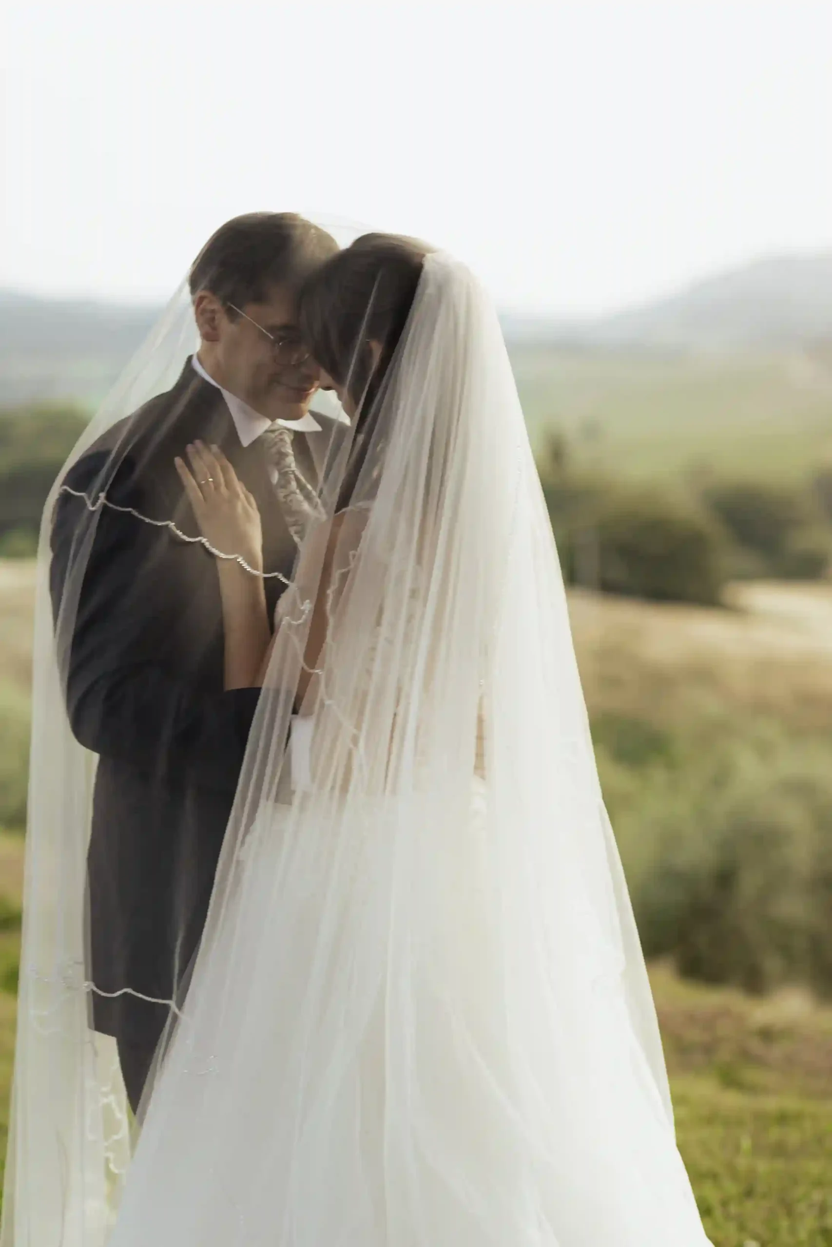 Brudepar står tett omslynget under brudesløret med utsikt over det toskanske landskapet. Et intimt øyeblikk fra et romantisk bryllup i Toscana. Foto av bryllupsfotograf Anna O. Brambilla