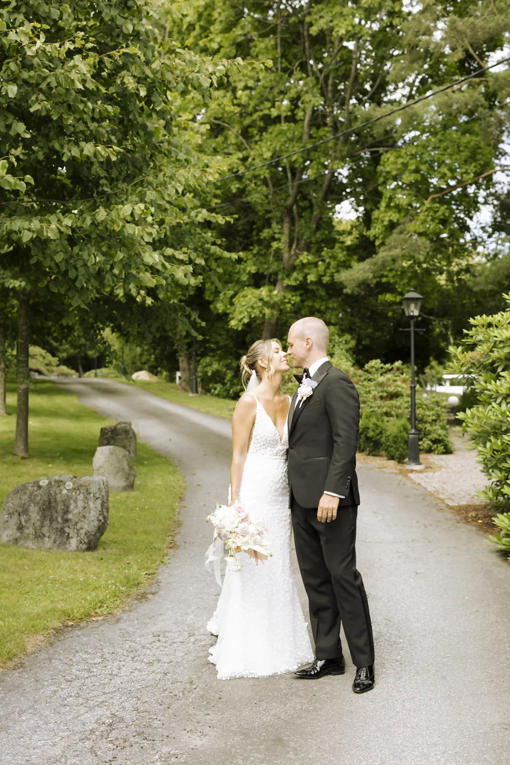 Brudepar fotograferes på en idyllisk landevei omgitt av grønt løvverk i Fevik. Bruden har på seg en elegant, tettsittende blondekjole og holder en lys bukett, mens brudgommen er kledd i klassisk sort smoking. Dette intime øyeblikket ble foreviget av Anna O. Brambilla, bryllupsfotograf Oslo, som også reiser for å dokumentere kjærlighet over hele Norge.