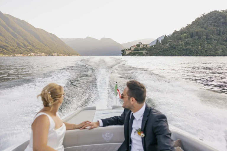 Bride and groom holding hands on a boat on Lake Como, leaving Villa del Balbianello in the distance during their elopement in Varenna. Photo by wedding photographer Anna O. Brambilla. Brudepar på båttur på Comosjøen med Villa del Balbianello i bakgrunnen etter elopement i Varenna. Foto av bryllupsfotograf Oslo Anna O. Brambilla.