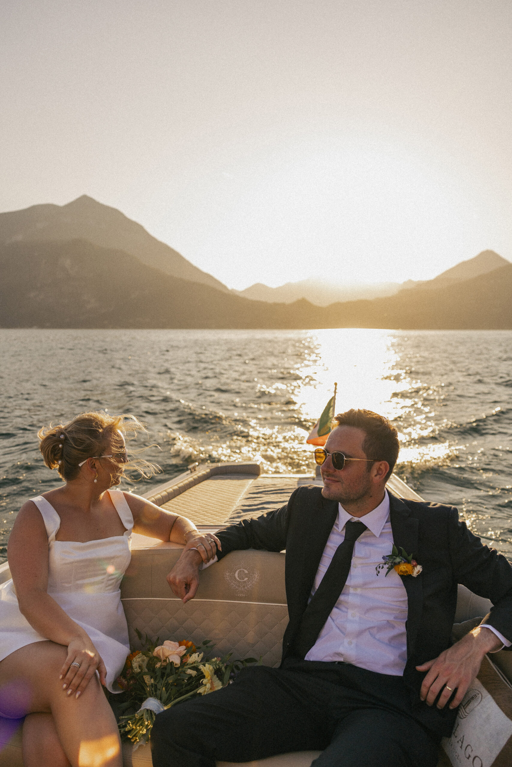 Bride and groom relaxing on a boat during golden hour on Lake Como, holding hands with bouquet nearby, captured during their elopement in Varenna. Photo by wedding photographer Anna O. Brambilla. Brudepar på båttur i solnedgang på Comosjøen, med blomsterbukett ved siden av. Foto fra elopement i Varenna av bryllupsfotograf Oslo Anna O. Brambilla.