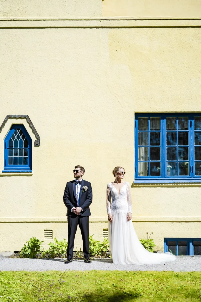 Modern wedding portrait of bride and groom standing against a yellow wall with blue windows in Molde – taken by Anna O. Brambilla, wedding photographer. Moderne bryllupsportrett av brudepar foran en gul vegg med blå vinduer i Molde – tatt av Anna O. Brambilla, bryllupsfotograf.